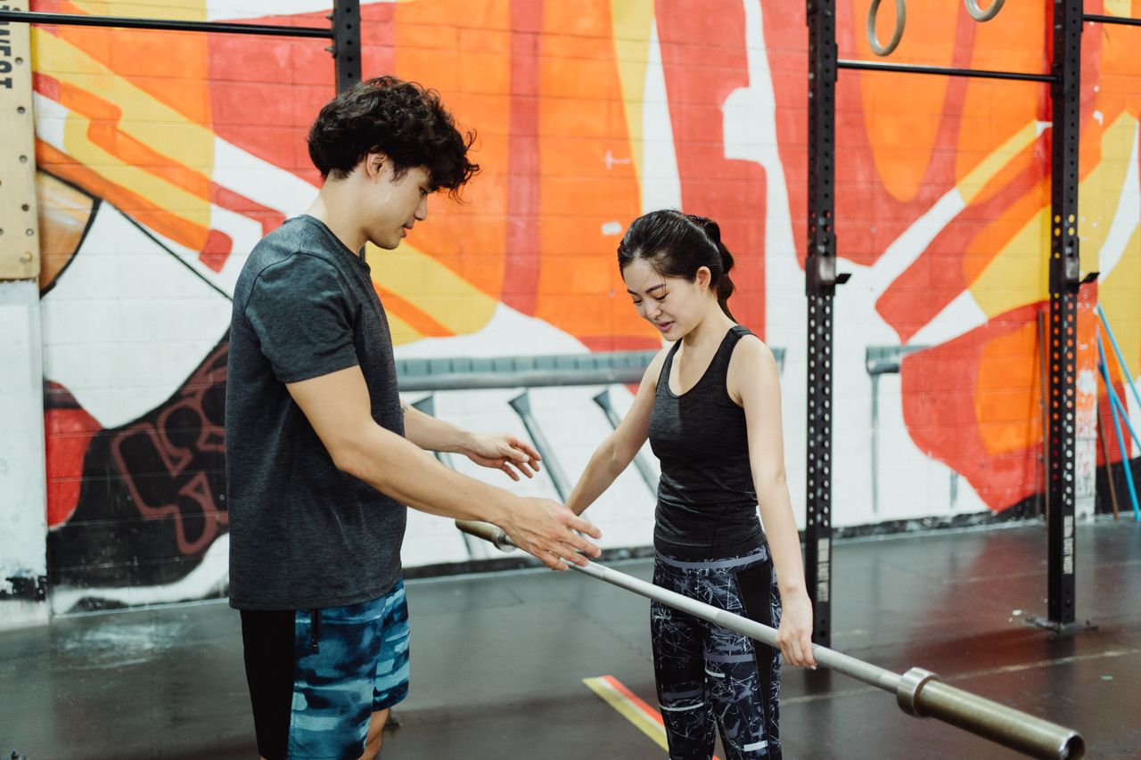 A male trainer instructing a female client on barbell lifting technique in a vibrant urban gym setting.